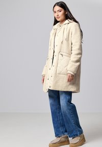 Mujer vestida con un abrigo de sherpa crema con bolsillos acolchados, jeans azules y zapatos de suela beige, de pie contra un fondo gris.
