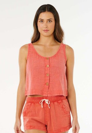 Frau trägt ein korallenfarbenes ärmelloses, geknöpftes Crop-Top und passende Shorts mit Kordelzug vor einem schlichten, hellen Hintergrund.