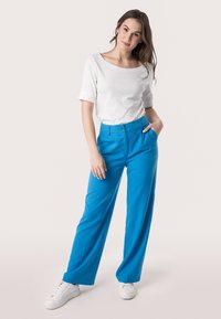 Witte kortarmige top gecombineerd met felblauwe wijde broek; heeft een frontale knoopsluiting en zakken, gestyled met witte sneakers.