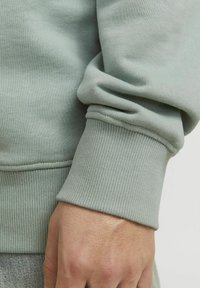 Sudadera verde claro con puños y dobladillo acanalados. Textura suave con un acabado liso. Mano reposando sobre la tela en una posición relajada.
