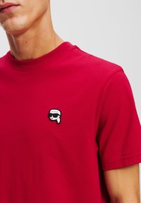 Tricou din bumbac roșu cu guler tip crew. Prezintă un mic patch brodat în alb și negru cu un personaj care poartă ochelari de soare pe piept.