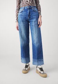 Los jeans de pierna ancha en azul claro presentan áreas desvanecidas y un dobladillo enrollado. Se combinan con zapatos estampados. El material parece ser mezclilla con una textura suave.