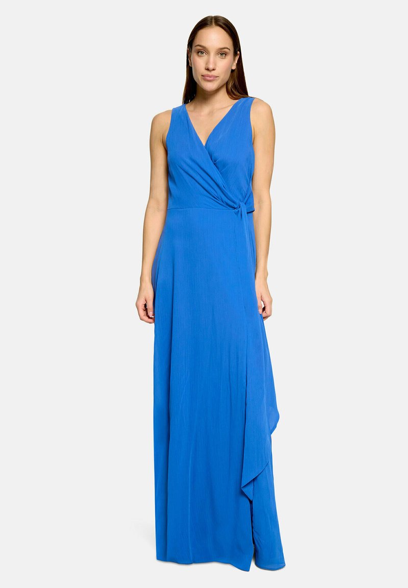 Femme portant une robe portefeuille sans manches, bleu vif, avec un col en V et un ourlet asymétrique, debout devant un fond gris clair uni.