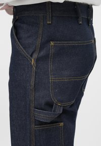 Jeans de ganga escura com costura amarela visível, apresentando vários bolsos, incluindo um bolso utilitário lateral, e um design de corte justo.