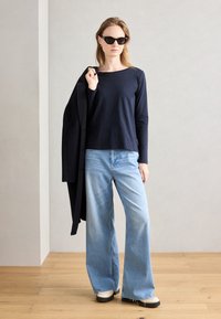 Schwarzes Langarmoberteil kombiniert mit hellblauen weiten Jeans. Das Model hält einen schwarzen Mantel und trägt schwarze Sonnenbrillen, während es auf einem Holzboden steht.