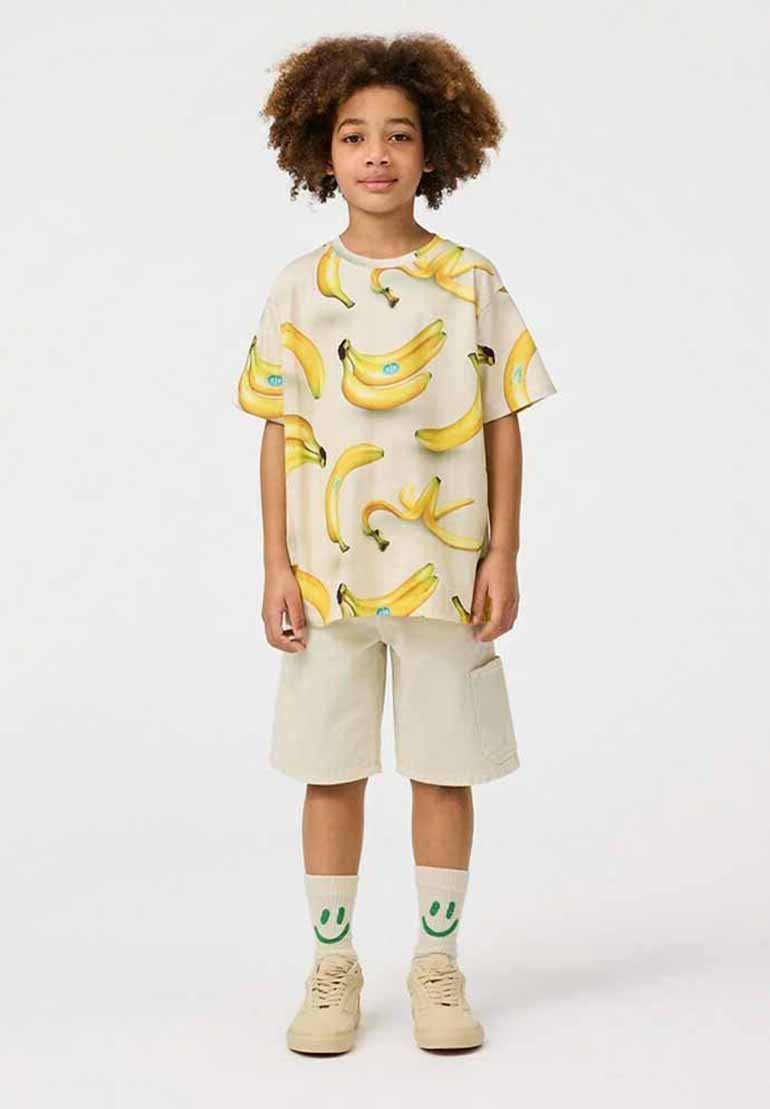 Lys beige t-shirt med et levende gult bananmønster, parret med beige shorts og cremede sneakers, og grønne smiley-face sokker.