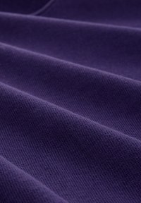 Lila Stoff mit einer strukturierten Oberfläche, die Falten aufweist, die ein Gefühl von Tiefe erzeugen. Das Material erscheint weich und deutet auf eine gestrickte oder gewebte Verarbeitung hin.