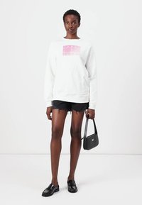 Witte sweatshirt met een roze grafisch logo, gecombineerd met zwarte verwaande shorts, een zwarte handtas en zwarte instappers.