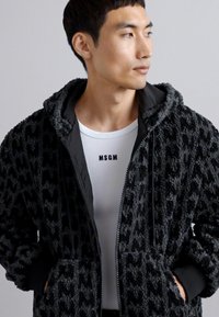 Black fleece jacket with a textured pattern, front pockets, and a hood. Worn over a white shirt featuring the text "MSGM."