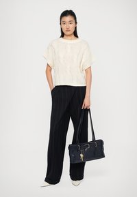 Cremfarbener Kabelflecht-Strickpullover, schwarze gestreifte weite Hose und eine marineblaue Ledertasche mit einem Schlüsselanhänger-Detail. Weiße spitze Schuhe.