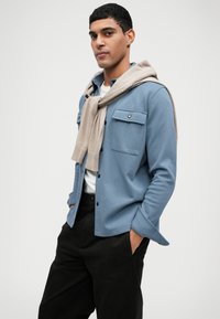 Světle modrá bunda na knoflíky s dvěma náprsními kapsami a béžovým pleteným šálkem volně položeným na ramenou; černé kalhoty.