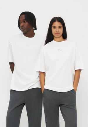 To personer iført hvide oversized t-shirts og mørkegrå bukser står side om side mod en ensfarvet baggrund.