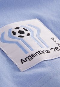 Tissu bleu clair avec un patch blanc comportant des éléments graphiques bleus et noirs, y compris un ballon de football et le texte "Argentine '78 TM FIFA."