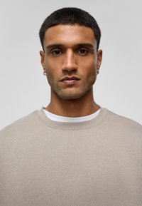 Licht beige sweatshirt met een ronde hals, eenvoudig ontwerp, zachte textuur en gelaagd met een witte onderhemd. Close-up portretweergave.