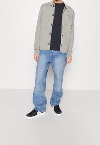 Grijze gebreide cardigan met zwarte knopen, gecombineerd met een zwart T-shirt en lichtblauwe losse spijkerbroek. Zwarte sneakers met witte accenten.