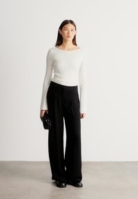 Hvit langermet topp med siksakmønster, kombinert med svarte bukser med vide ben og svarte sko, holder en liten svart veske.