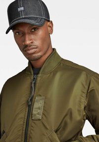 Veste bombardier vert olive avec fermeture éclair et poches latérales, associée à une casquette en denim foncé avec des coutures contrastantes sur la visière.