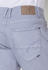 Gai zila džinsa bikses aizmugures kabata ar brūna ādas zīmola uzlīmi un redzamām jostu cilpām, šuvēm un kniedēm.