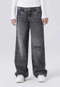 Široke sive traperice s izblijedjelom teksturom, sa istrošenim koljenom i klasičnim dizajnom s pet džepova. Uparene s bijelim tenisicama.