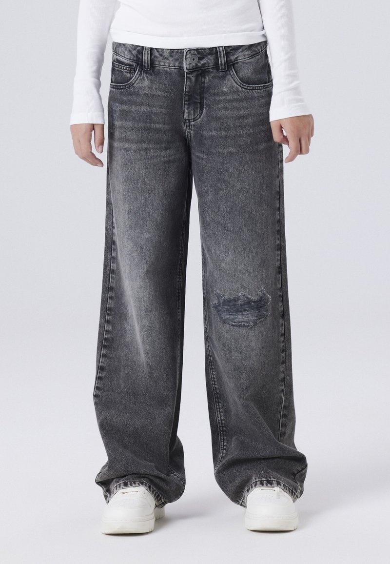 Široke sive traperice s izblijedjelom teksturom, sa istrošenim koljenom i klasičnim dizajnom s pet džepova. Uparene s bijelim tenisicama.