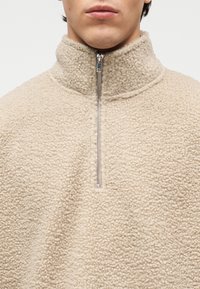 Μπεζ φλις πουλόβερ με ψηλό γιακά, μπροστινό μέρος με μισό φερμουάρ και υφή αφράτης, βελούδινης επιφάνειας. Διαθέτει μεταλλικό φερμουάρ.