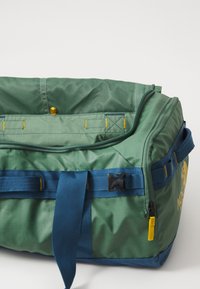 Grønn og blå duffelbag med en teksturert overflate, mesh-topp, gule detaljer og en svart spenne. Har slitesterkt stoff og justerbare stropper.