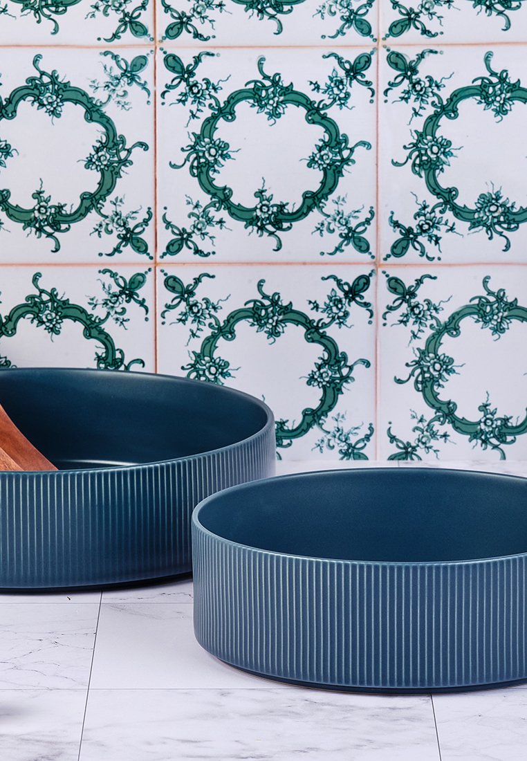 Zwei runde blaue Keramikschalen mit vertikaler Rillenstruktur, vor einem grün-weißen gemusterten Fliesenhintergrund.