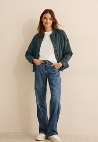 Giacca bomber verde scuro, t-shirt bianca, jeans blu a vita alta e sneakers bianche; modella in piedi con le mani in tasca su uno sfondo neutro.