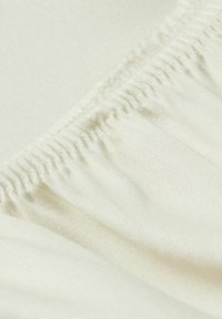 Crème-kleurige stof met een gladde, iets glanzende textuur. Heeft naar binnen toe verzamelde randen en subtiele stiksels langs de naad.