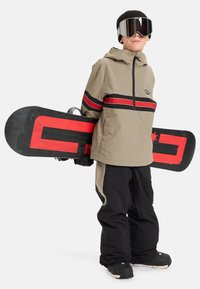 Kind in beige en zwarte sneeuwuitrusting houdt een zwarte snowboard met rode ontwerp vast, draagt een zwarte beanie en gespiegeld skibril.