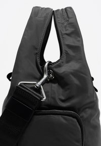 Črna najlon torba z teksturirano površino, opremljena s srebrnim kovinskim zatičem in tkanim trakom, poudarjena s čistimi, minimalnimi oblikovalskimi elementi.