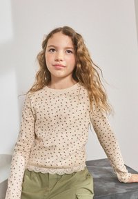 Un pull beige à manches longues avec un motif floral, une finition en dentelle au bas, et une variation de texture. Associé à un pantalon cargo vert clair.