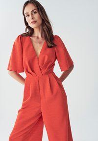 Joven mujer con cabello largo castaño, vistiendo un mono texturizado de un brillante color naranja con mangas anchas y un escote pronunciado, de pie contra un fondo claro.