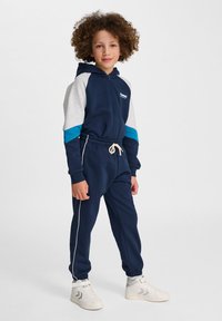 Sjöfararträningsdräkt med zip-up hoodie, grå och teal detaljer. Innehåller elasticitet i midjan och ärmarna, med snörning, parat med vita sneakers.