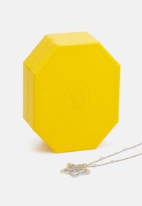 Osemhranná žltá šperkovnica s textúrovaným povrchom, s logom labute, vedľa strieborného náhrdelníka s hviezdou a žltými akcentmi.