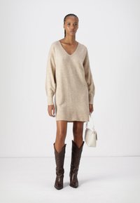 Beige kötött ruha V-nyakkal, hosszú ujjakkal, bordázott aljjal, barna bőrcsizmával, cowboy stílusban, valamint egy kis fehér táskával.