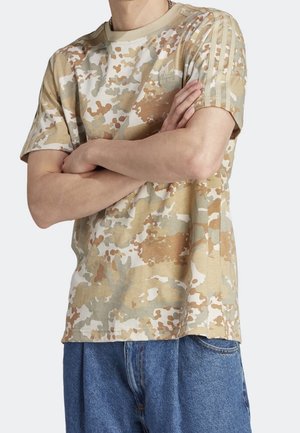 Personne les bras croisés portant un t-shirt Adidas camouflage beige et marron clair ainsi qu'un jean denim bleu, sur un fond clair uni.