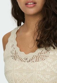 Camiseta sin mangas de punto en color blanco roto con escote festoneado, que presenta intrincados patrones de encaje y costuras texturizadas.