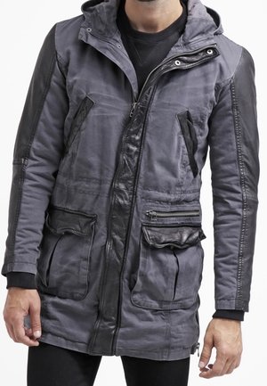 Parka grise pour homme avec capuche, comprenant des accents en cuir noir, des poches zippées et une finition en tissu texturé. Manches longues avec poignets côtelés.