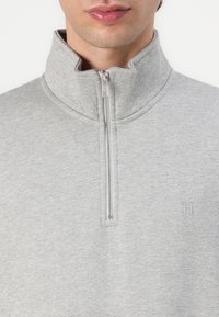 Sivi zip-up dukserica izrađena od mekog, teksturiranog materijala ima visoki ovratnik i suptilan logo na grudima. Zahvaljujući metalnoj sivi zatvaraču.