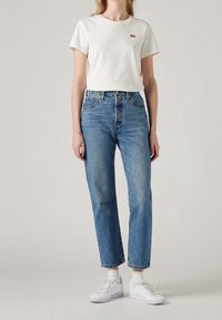 Camiseta blanca de manga corta con un pequeño logo rojo, combinada con vaqueros de mezclilla azul de cintura alta y zapatillas blancas con suelas texturizadas.