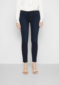 Jeans skinny de color azul oscuro con una cintura alta, presentando una textura de denim suave y detalles de costura sutiles, combinados con sandalias deslizables beige.