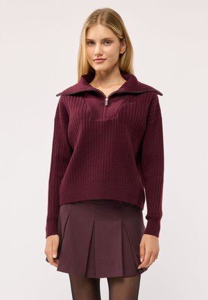 Giovane donna che indossa un maglione a coste color granata con mezza cerniera e una gonna plissettata granata su uno sfondo semplice.
