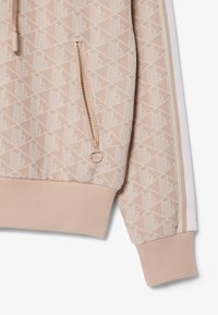 Beigefarbener Jackenärmel mit weißem geometrischem Muster, beige Reißverschlusstasche, geripptem Bündchen und weiß-beige gestreiftem Seitenpaneel.