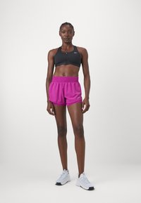 Černá sportovní podprsenka s předním zipem, spárovaná s růžovými šortkami s vysokým pasem a elastickým pasem. Bílé sportovní boty s minimalistickým designem.
