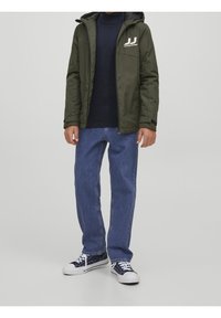 Bambino che indossa una giacca a cappuccio verde oliva, un maglione blu scuro, jeans blu e scarpe nere e bianche, in piedi contro uno sfondo neutro.