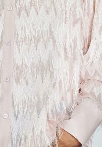 Lichtroze blouse met een zigzagpatroon en lange franjes. Het heeft een knoopsluiting aan de voorkant en een gladde stofstructuur.