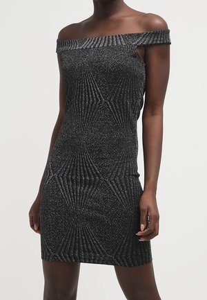 Kvinde iført en sort figursyet off-shoulder kjole med et geometrisk sølvglimmermønster, stående foran en ensfarvet baggrund.