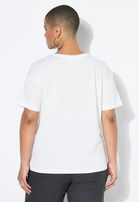 Personne avec des cheveux courts et des boucles d'oreilles créoles portant une chemise blanche à manches courtes simple et un pantalon sombre, faisant face à l'opposé de l'appareil photo.