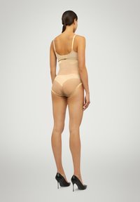 Beige shapewear bodysuit med justerbara band och en transparent ryggdesign, kombinerad med svarta högklackade skor. Slät textur och åtsittande form.